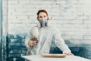 Woman in protective suit and gas mask using spray gun indoors against a painted brick wall.